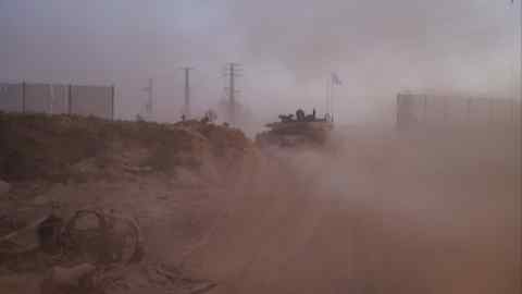 Seen through clouds of churned-up dust, a tank speeds away through a breach in a wire fence