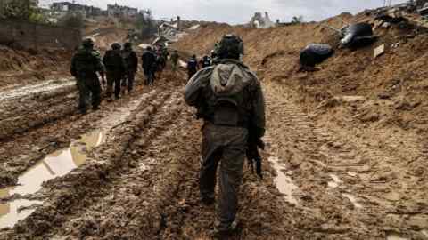 A group of armed soldiers wearing tactical gear walk along a muddy, war-torn path surrounded by destroyed buildings and debris