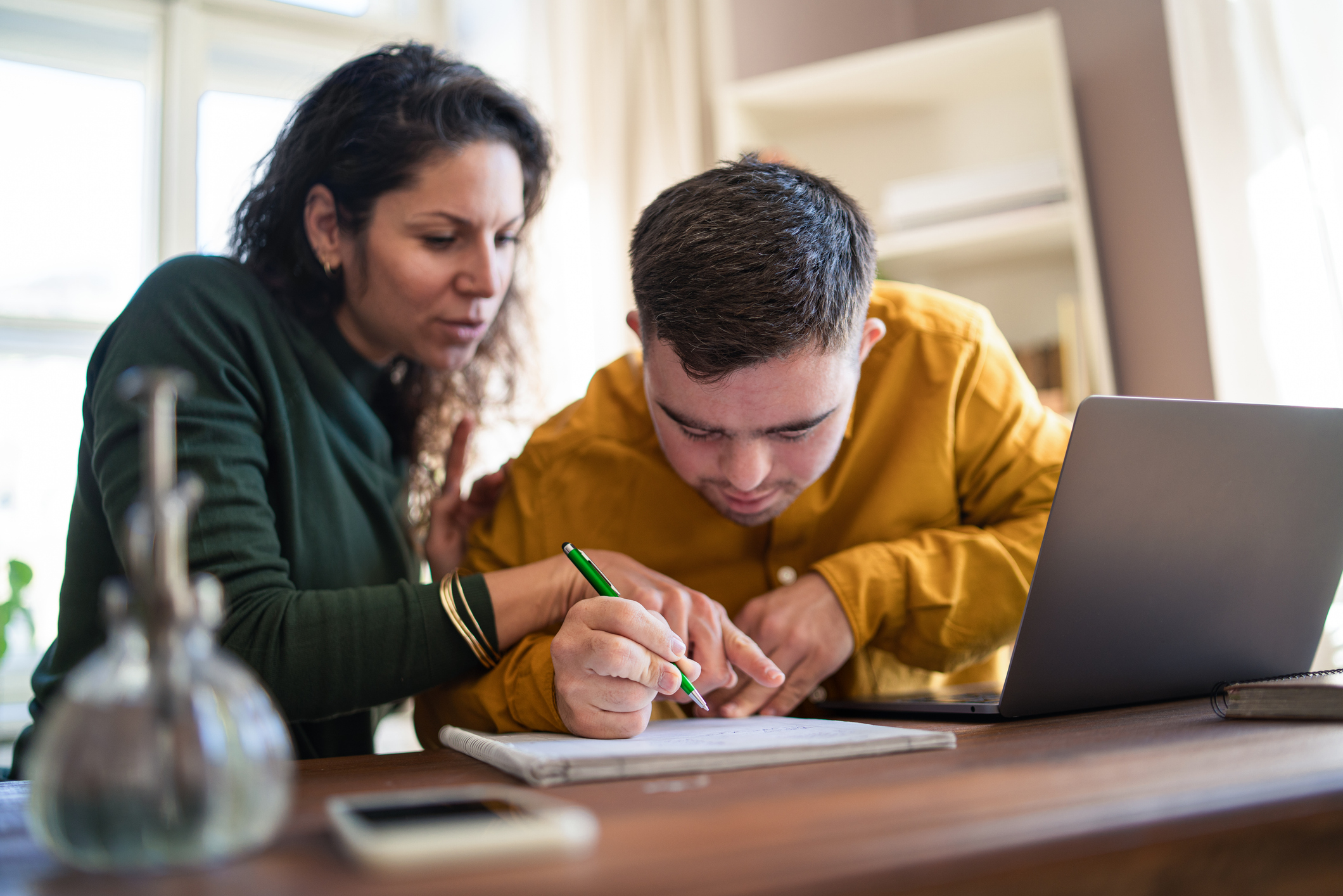 Mid adult woman helping young man with Down syndrome with learning and doing homework online on laptop.