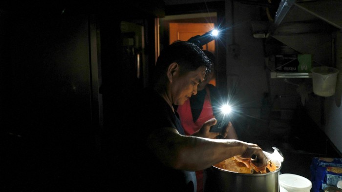 A cook works in a dimly lit kitchen during a power outage in Madrid, using two mobile phone flashlights to illuminate his workspace while stirring a large pot.