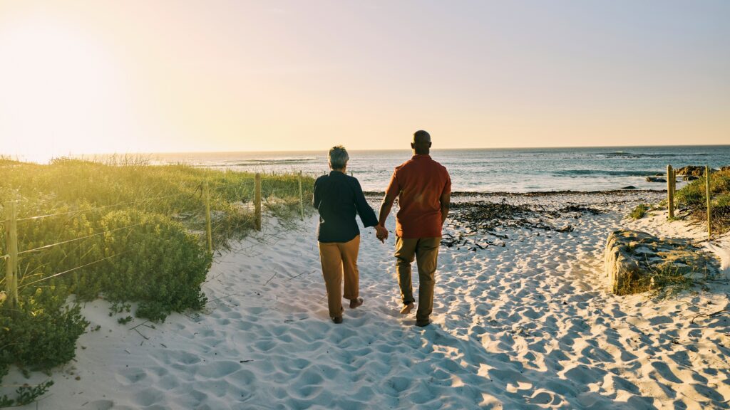 An older couple walk toward the beach sunset, holding hands.