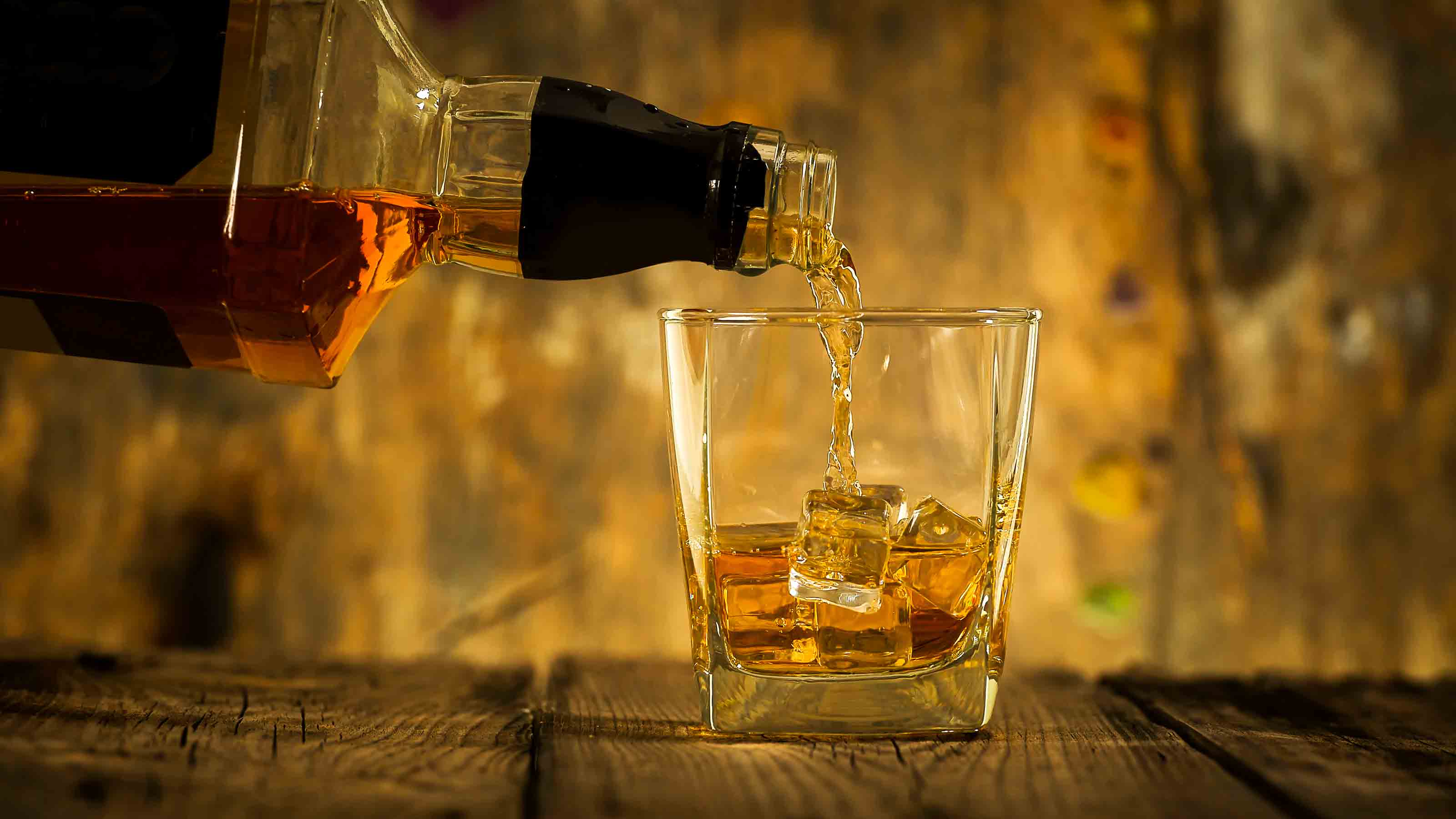 Barman pouring whiskey in front of whiskey glass and bottles on black table with whiskey and ice on a glass table