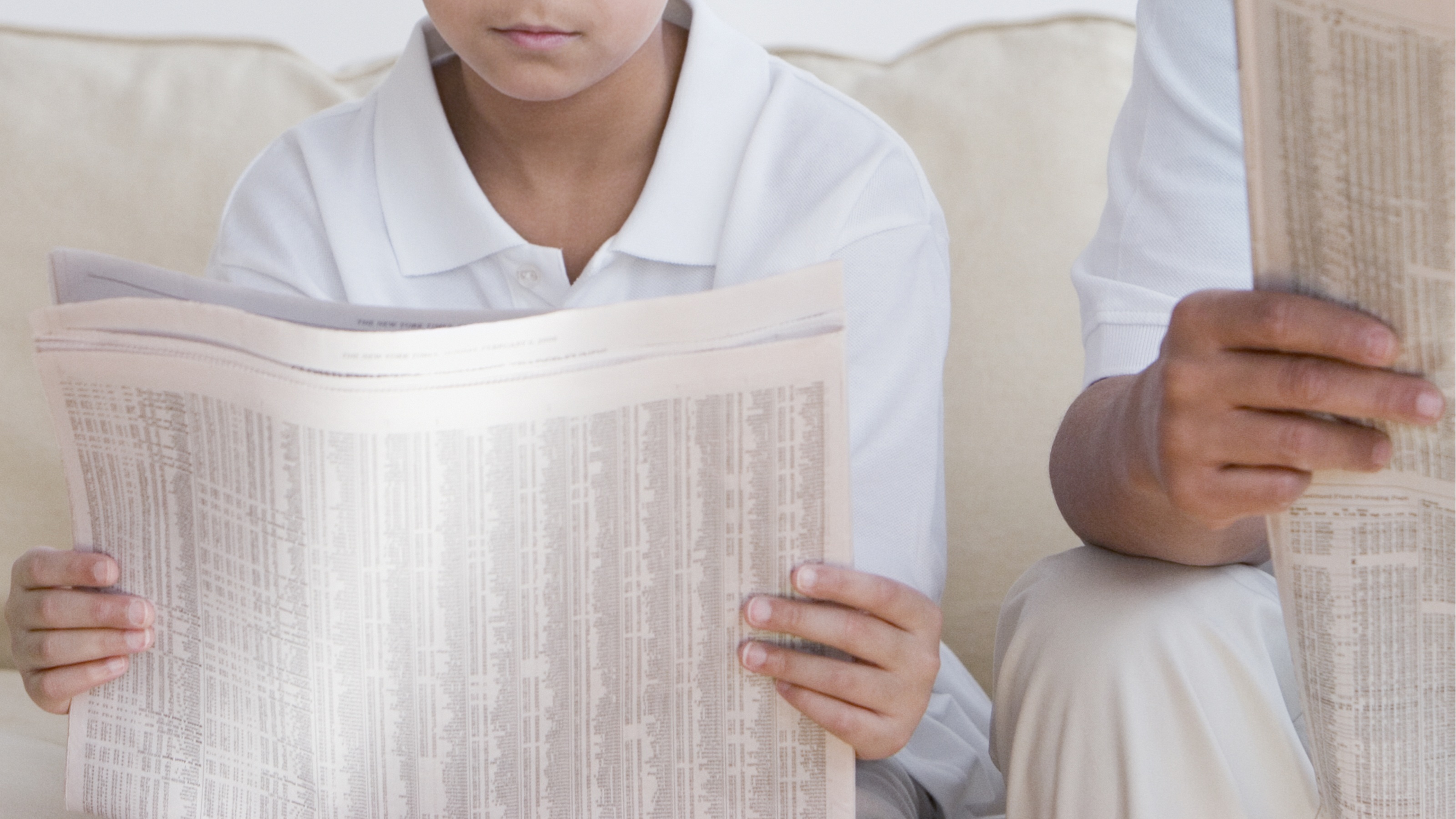 A young boy and his father read the newspaper side by side on the sofa.