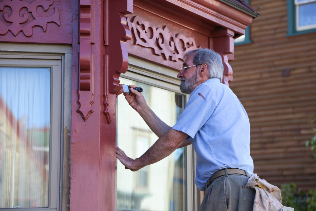 An older man in an historic town paints his house, with careful attention to detail, while standing on a ladder.