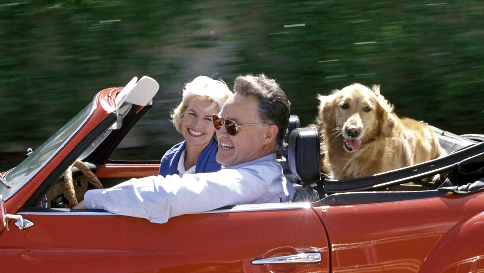 A retired couple drive in a convertible with a dog in the back seat.