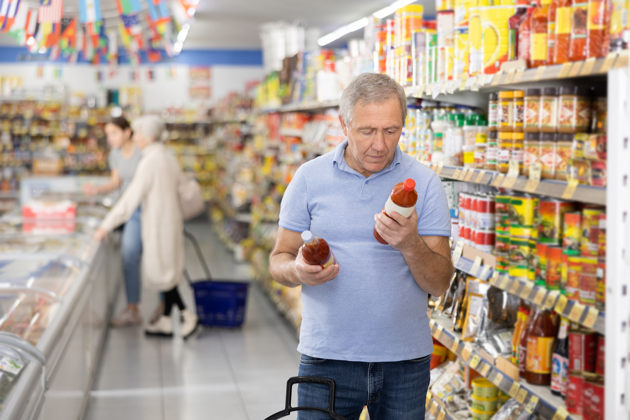 A mature man is reading the labels to compare different brands of hot sauce inside a grocery store.