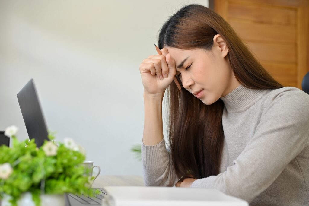 Side view of a stressed young woman sitting at a desk with a laptop, her eyes closed and brow furrowed as she presses a pencil against her forehead in frustration. This visualizes the overwhelming academic pressure, financial hardship, and mental health struggles that contribute to the high college dropout rate, where nearly 40% of students leave without earning a degree. Source: The College Investor