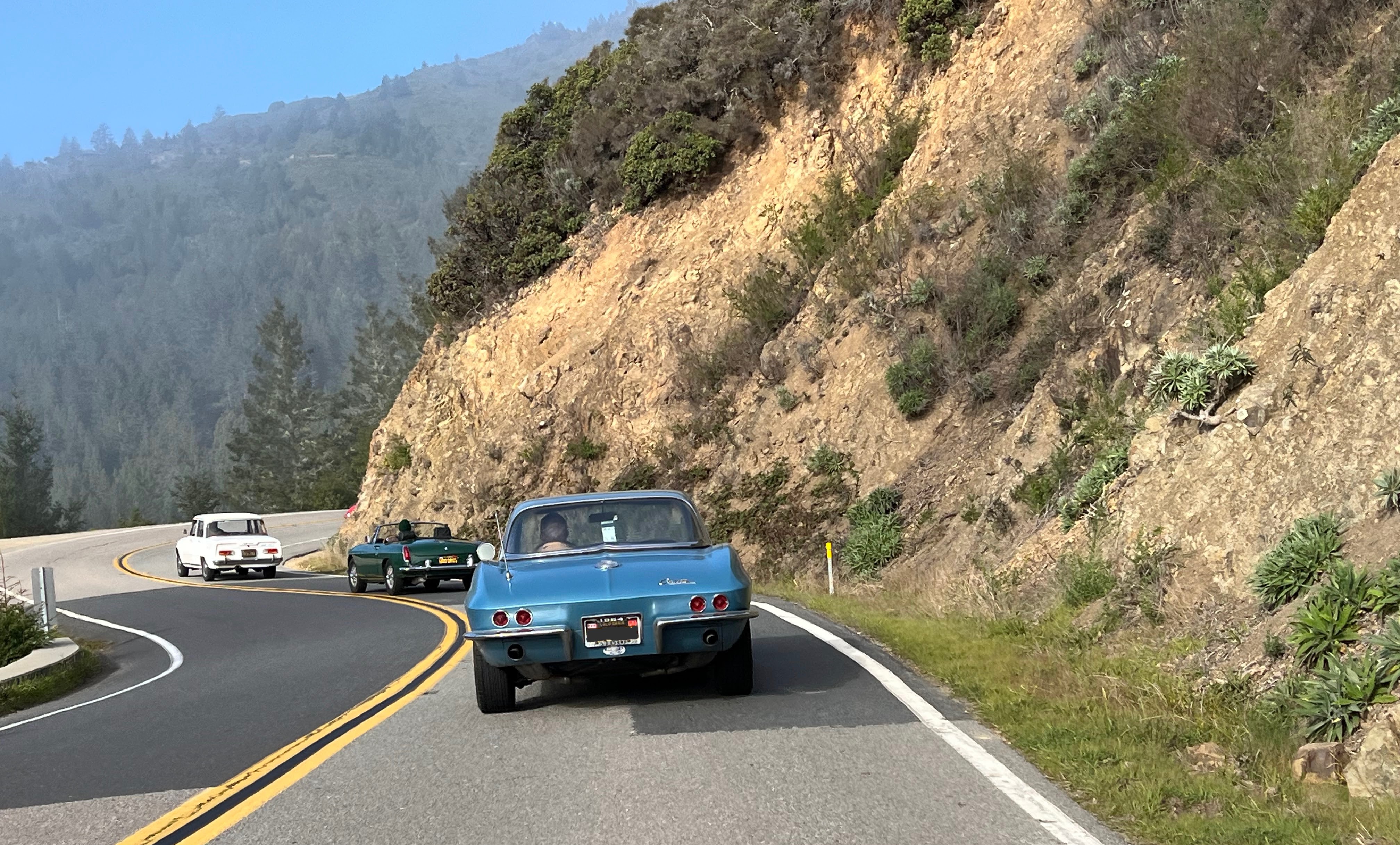 Three classic cars wind their way along California Bay Area roads with a view of mountains.