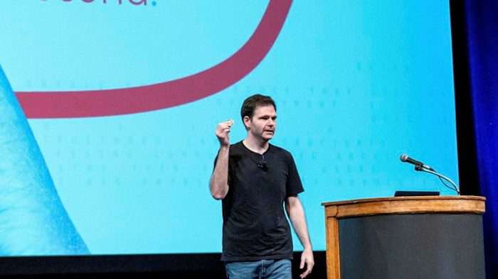 Jonathan Ross stands on stage speaking and gesturing with one hand raised at the GenAI Summit, with a microphone and podium nearby.