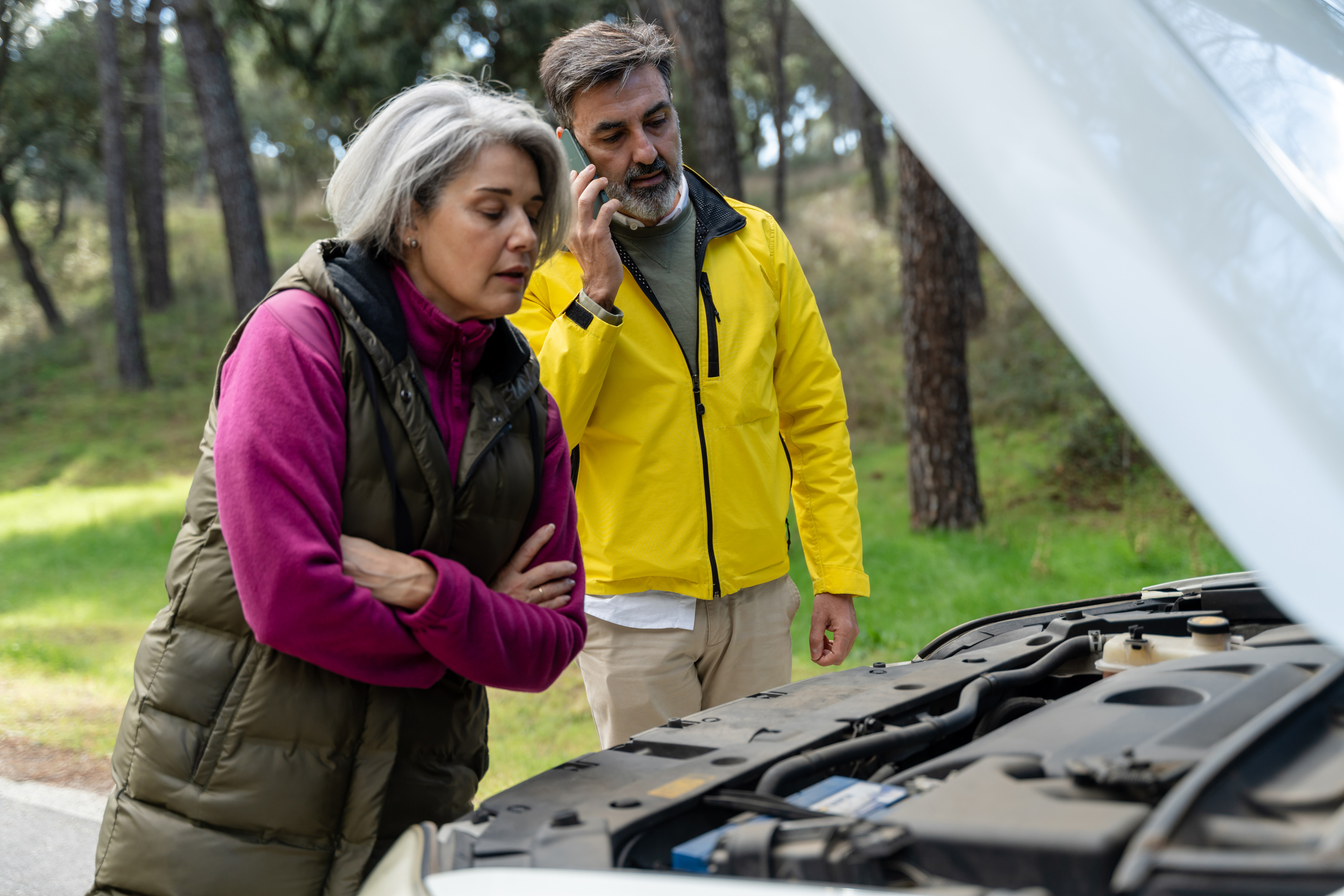 A mature couple experiencing car trouble inspects the engine on the side of the road.