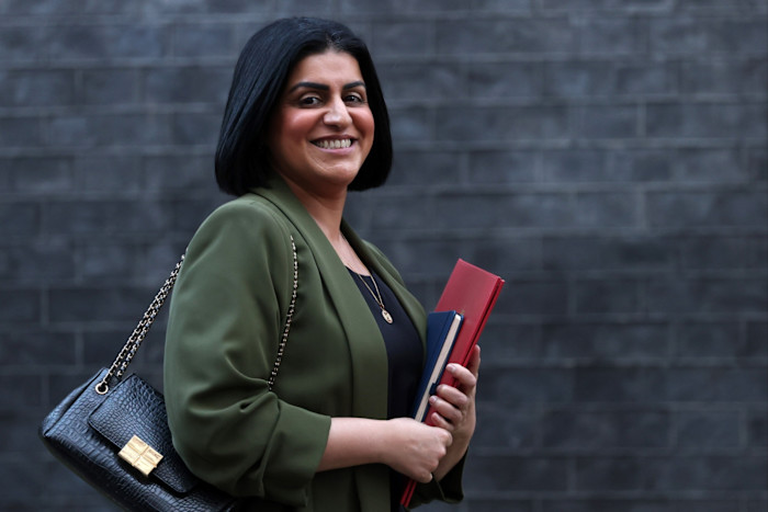 Shabana Mahmood smiles while holding documents and leaving a building, wearing a green blazer and carrying a black handbag