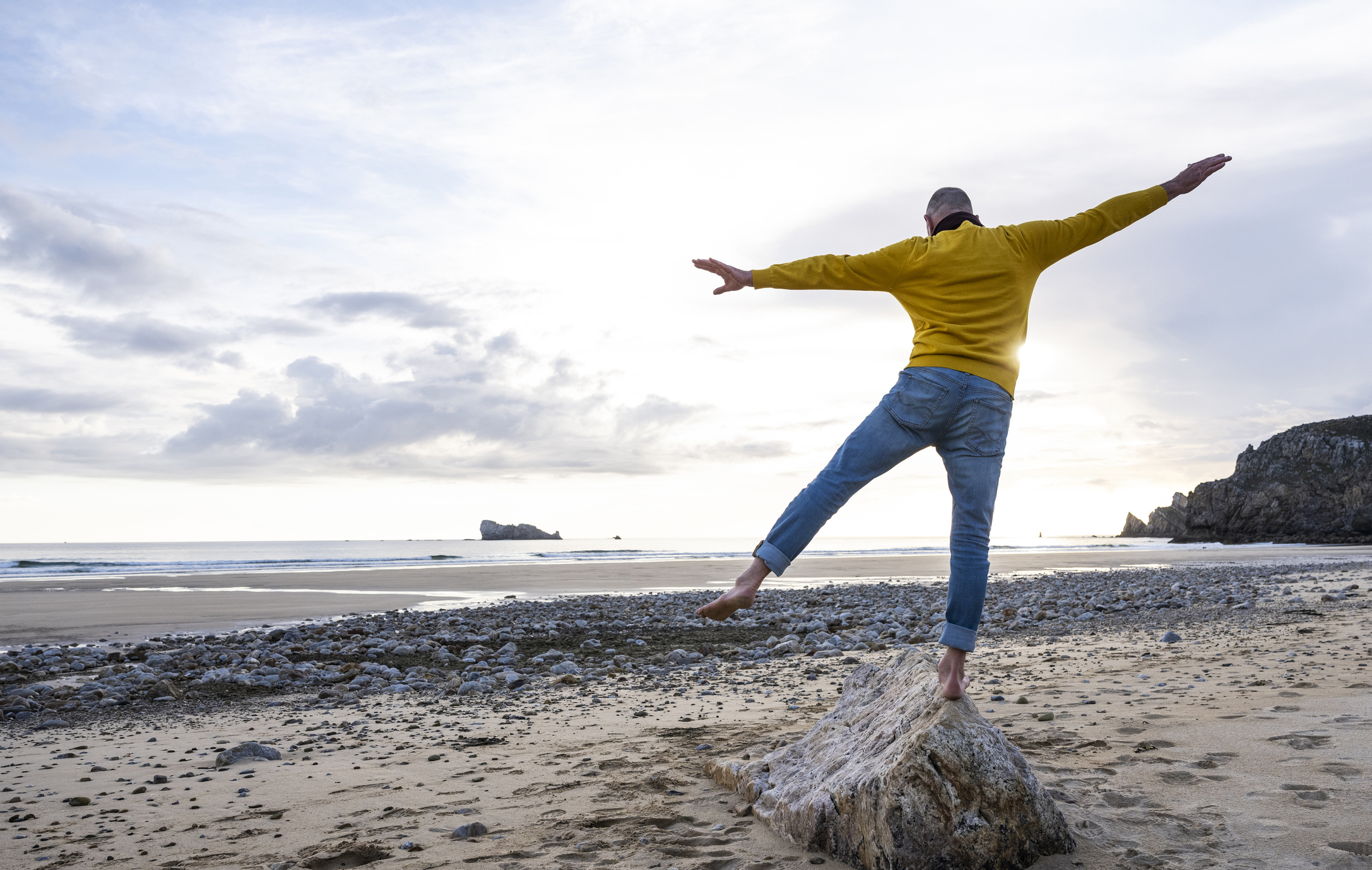 An older man balances on a rock on the beach, wearing jeans and a yellow long-sleeve shirt.