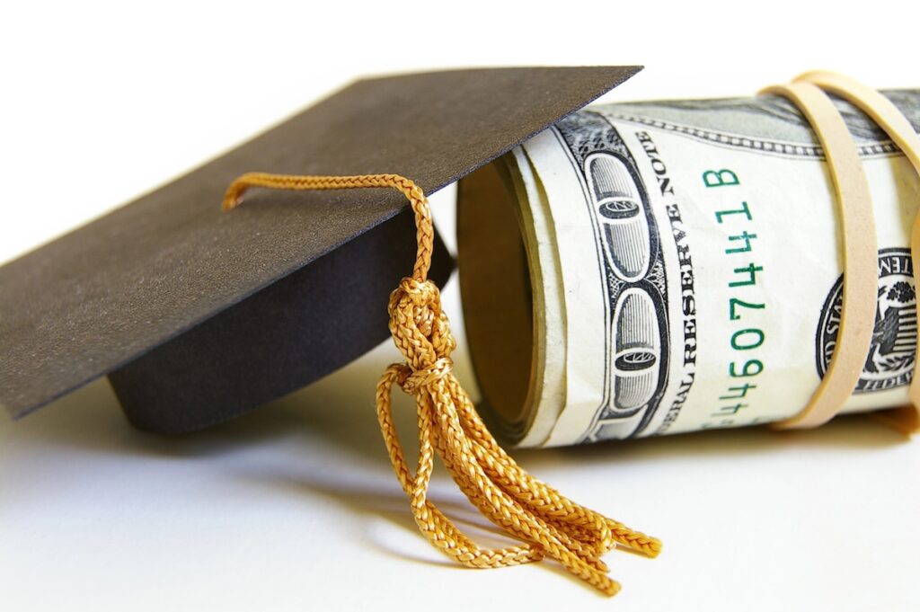 Close-up of a black graduation cap with a gold tassel resting next to a thick roll of U.S. dollar bills secured by a rubber band. This image symbolizes the significant financial shifts facing students and families in 2026, including the end of Grad PLUS loans, new caps on Parent PLUS borrowing, and the transition to the Repayment Assistance Plan (RAP). Source: The College Investor