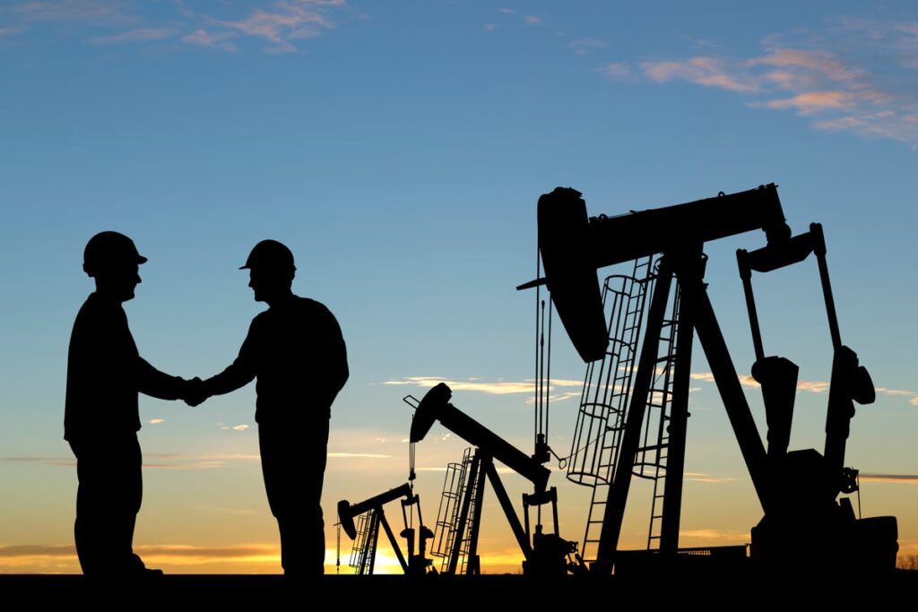 In front of a series of oil rigs, two workers shake hands.