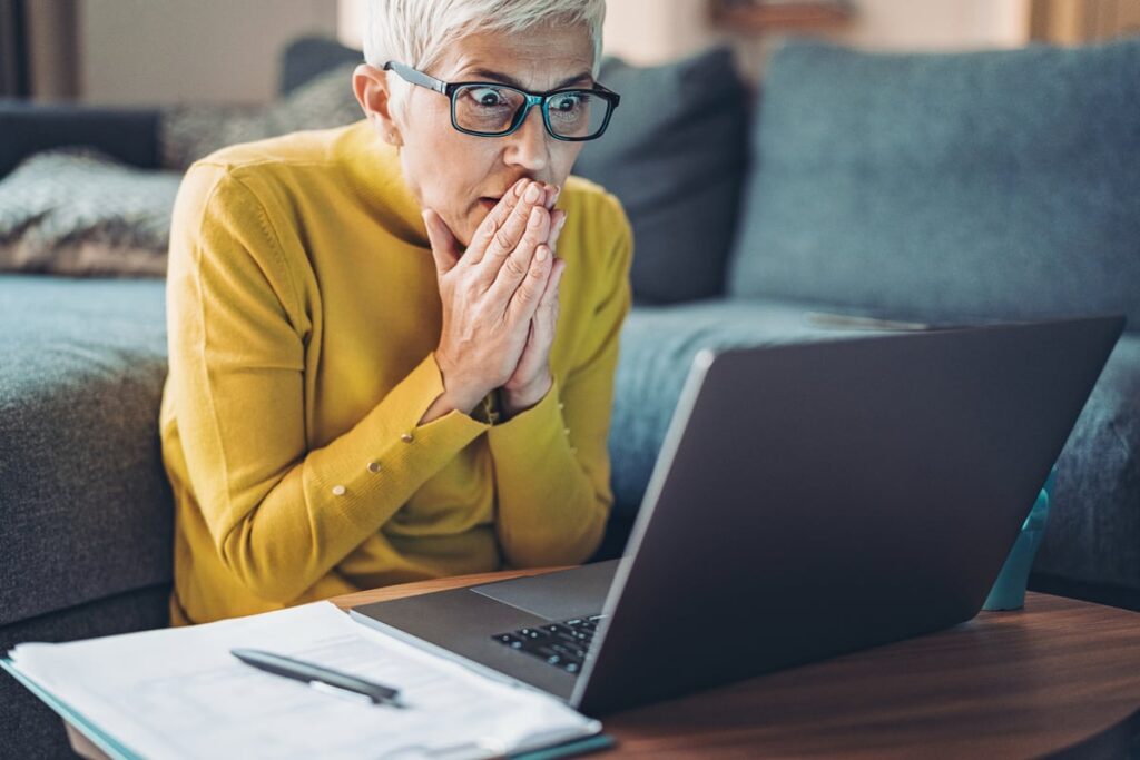 A shocked person staring at a laptop with their hands over their mouth.