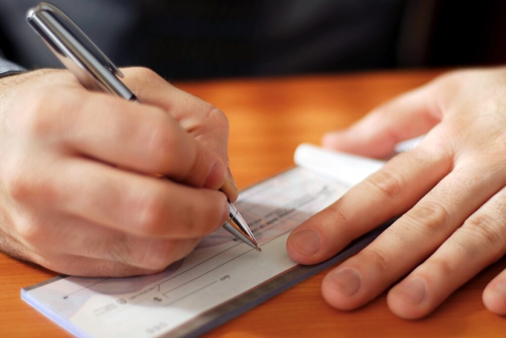 Close-up of a person's hands writing a check on a wooden desk. This image illustrates the reality of out-of-pocket college costs, where most families end up paying between $25,000 and $100,000 for a bachelor's degree after financial aid is applied. Source: The College Investor