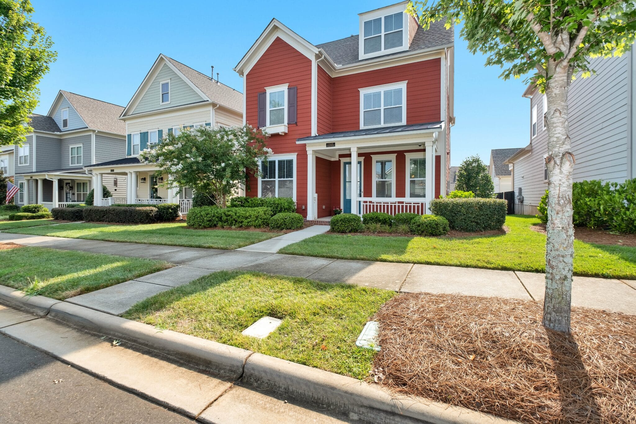 This image showcases a charming two-story red house with white trim and a welcoming front porch, situated in a well-maintained suburban neighborhood. Lush green lawns, manicured bushes, and mature trees line the sidewalk and street, creating a pleasant residential atmosphere. Other similar homes, some in lighter shades of gray and beige, are visible further down the street under a clear blue sky. This visual context is highly relevant to an article discussing reverse mortgages, home equity loans, and HELOCs, as it depicts the type of residential property where homeowners, particularly those 62 or older, might consider leveraging their home equity to supplement their monthly retirement income. The house represents the valuable asset that serves as collateral for a reverse mortgage, highlighting the importance of property ownership for seniors seeking financial flexibility.