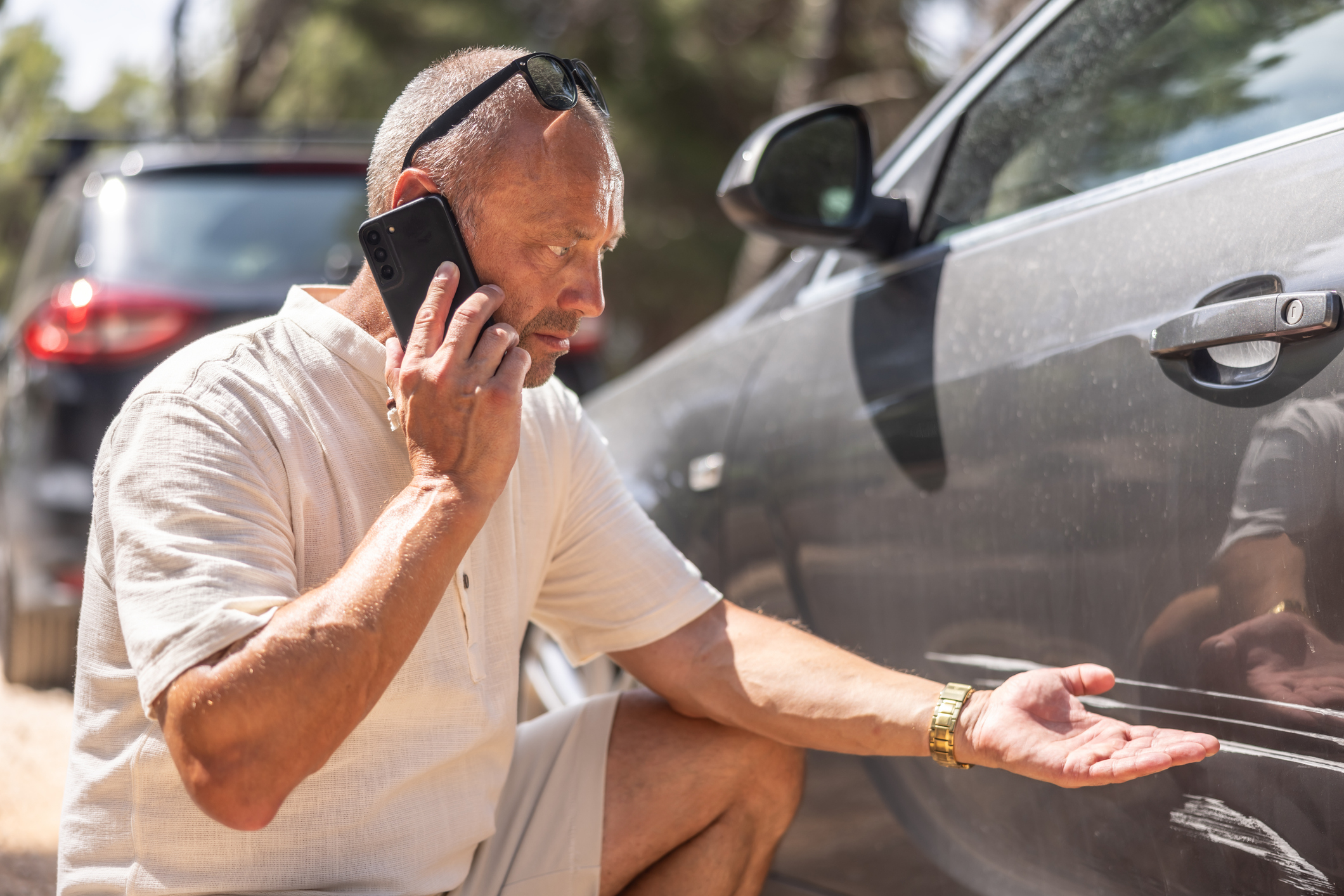 A man inspects damage on his car while calling car insurance.