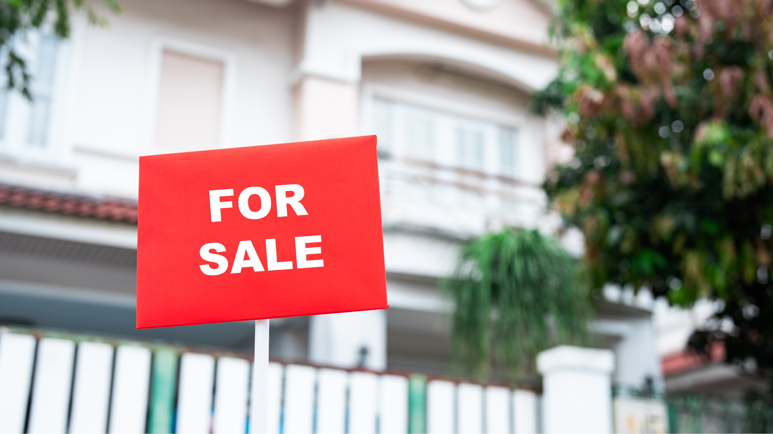 A "For Sale" sign in front of a home with a white fence.