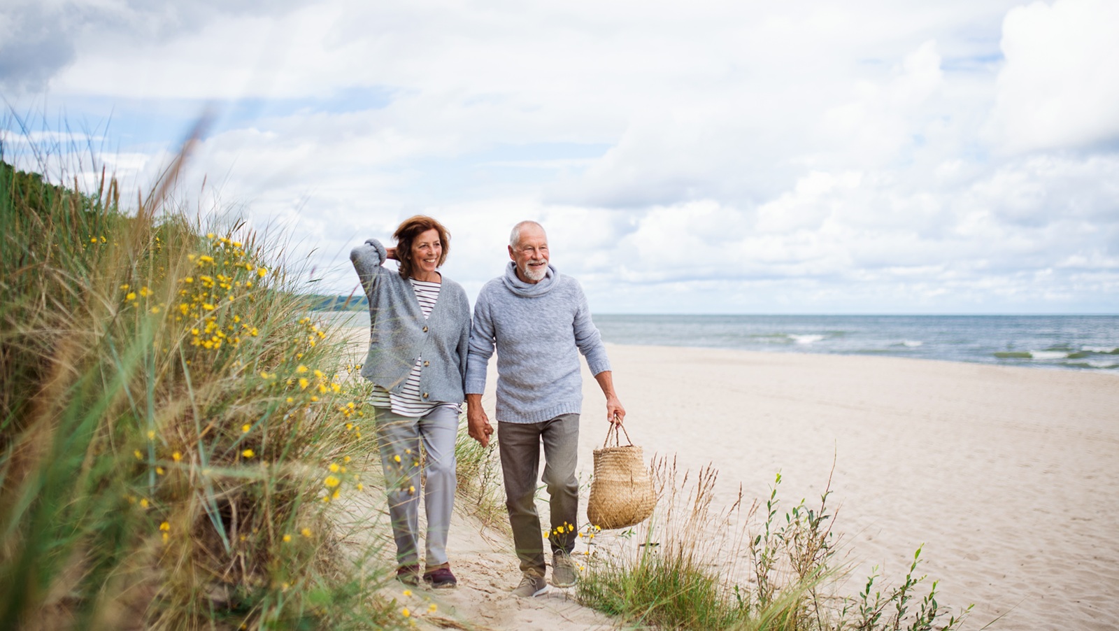 Happy senior woman and man holding hands and walking outdoors on sandy beach in autumn.
