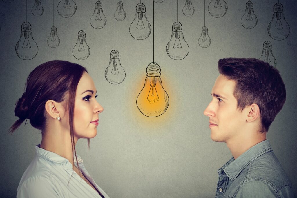 Profile views of a young woman and a young man facing each other, set against a grey background adorned with hand-drawn lightbulbs. A single bright yellow lightbulb glows centrally between them, symbolizing the realization or "bright idea" regarding the shifting gender dynamics in higher education. This visual metaphor accompanies an analysis of the growing gender gap in college degree attainment, where women now outpace men in earning Associate's, Bachelor's, Master's, and Doctoral degrees. Source: The College Investor