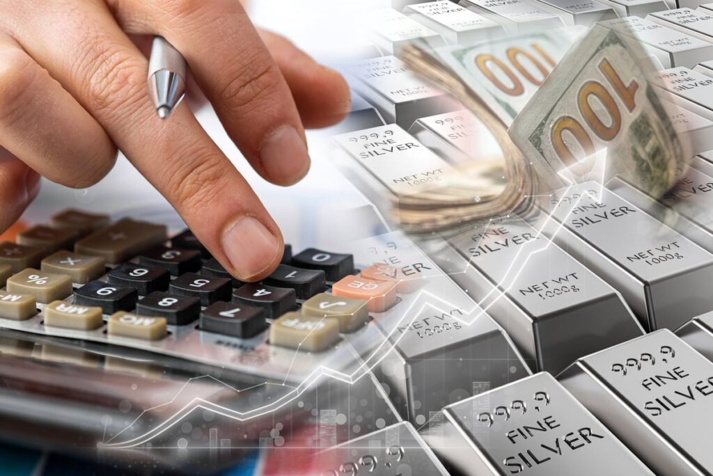 A hand using a calculator with money and silver bars in the background.