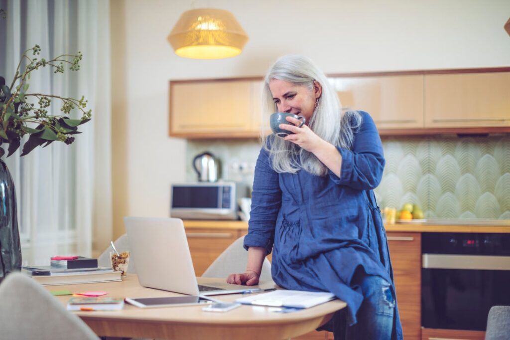 Person sipping coffee while standing in front of a desk and looking down at a laptop.