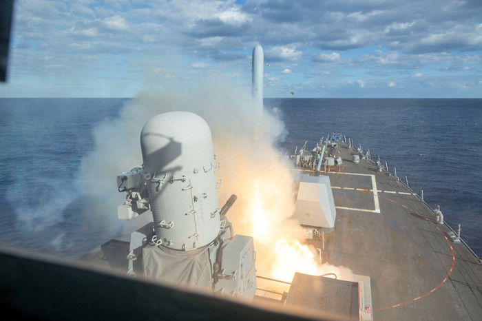 A rocket launches from a warship at sea, with smoke and flames visible on the ship’s deck under a partly cloudy sky.