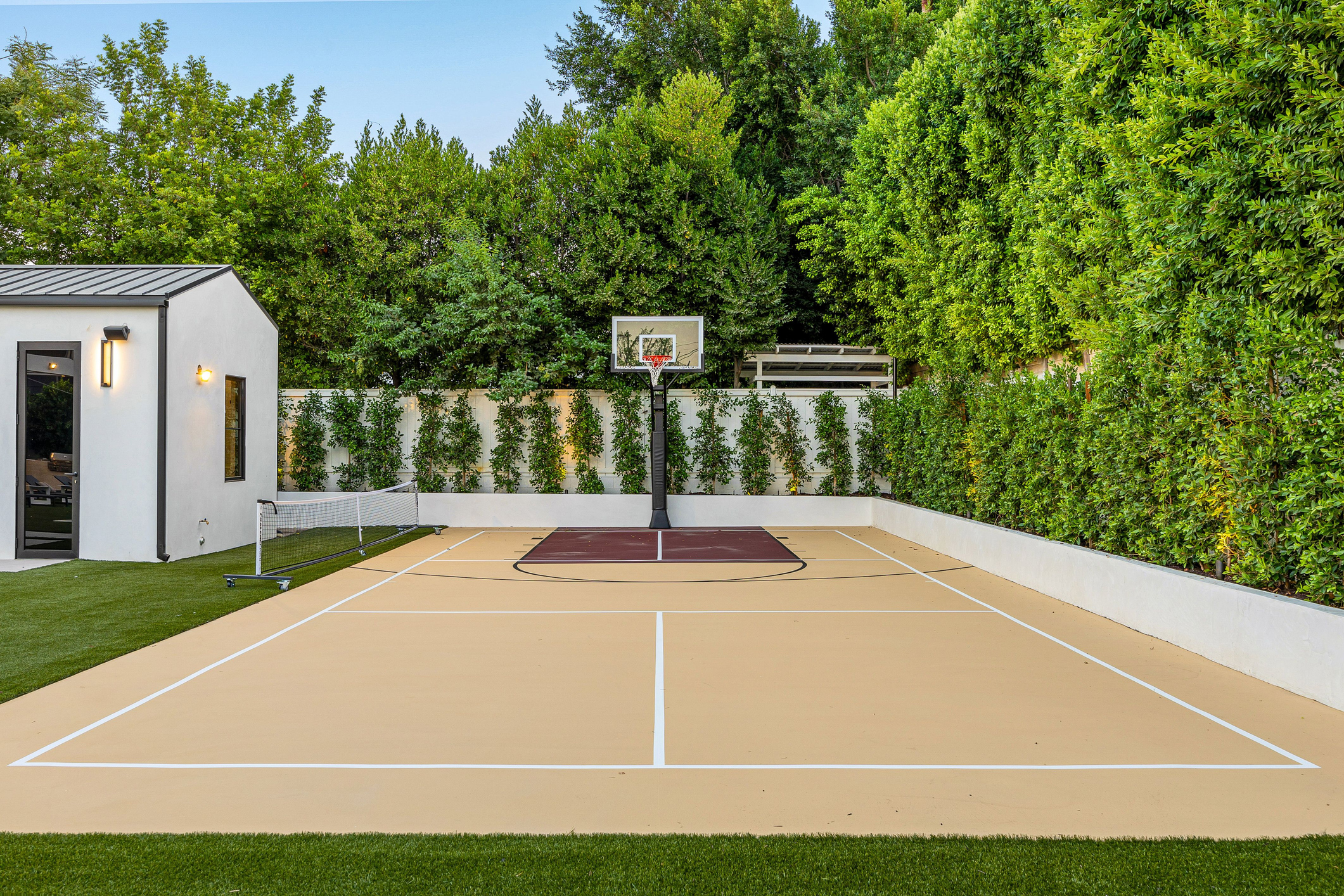 Modern backyard sports court with basketball hoop and tennis net surrounded by lush greenery.