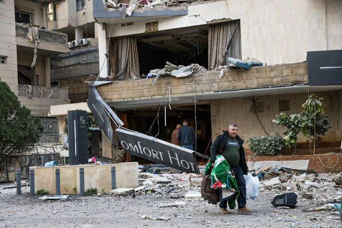 A man carrying bags and a blanket walks away from a heavily damaged hotel with debris scattered outside after an airstrike.