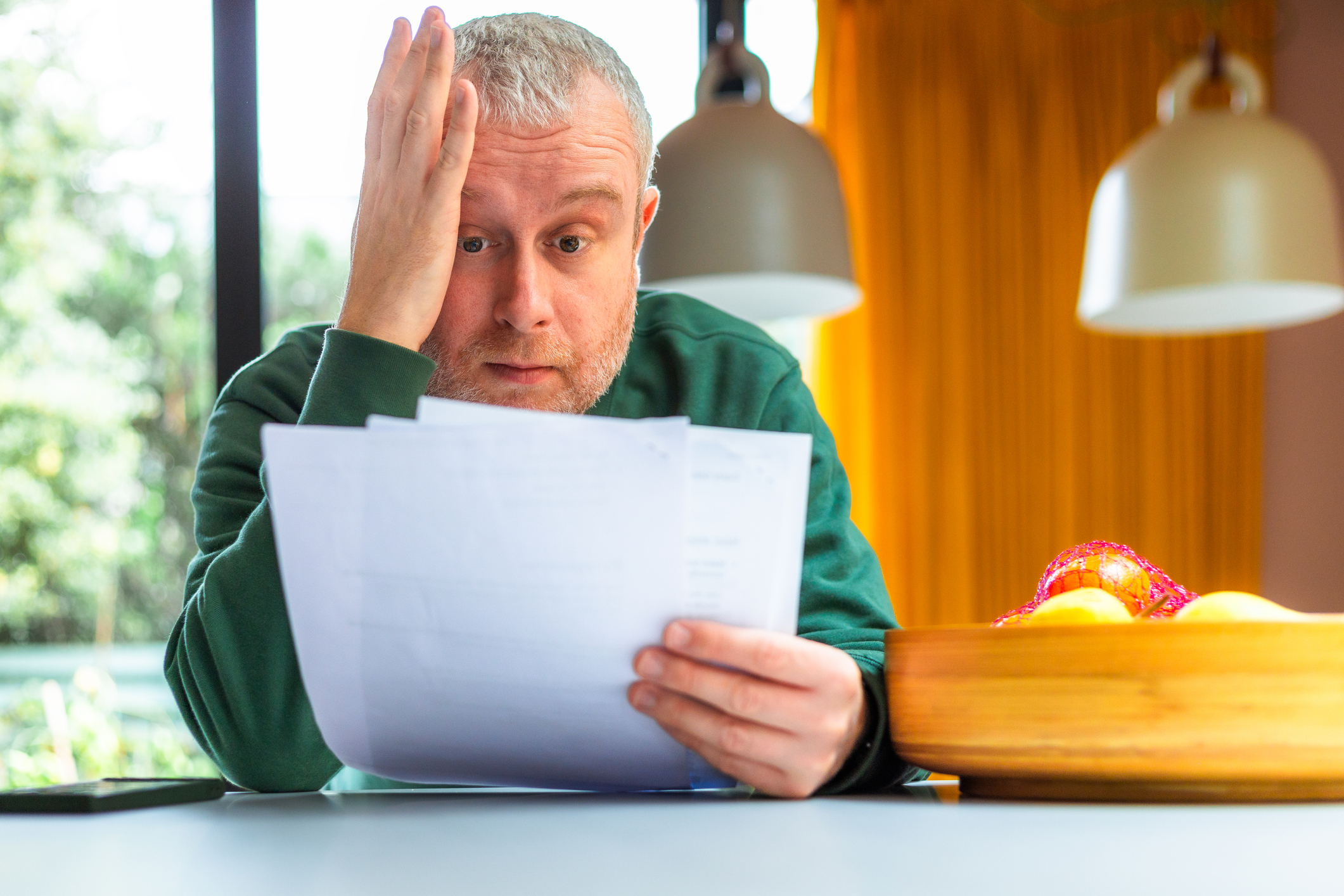 Portrait of a mid adult man checking his energy bills at home. He has a worried expression and touches his face with his hand while looking at the bills. He is sitting in his kitchen at home.