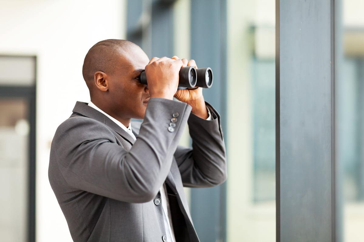 An investor looking into the distance through binoculars while facing an office window. 