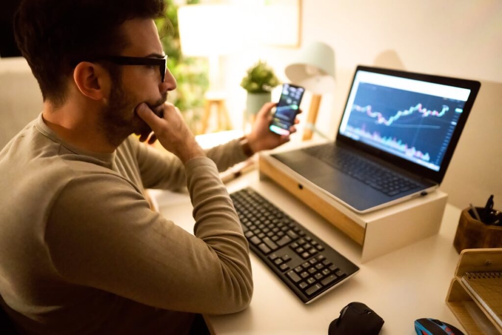 Person sitting at a desk in front of laptop while holding cell phone and looking at stock chart.