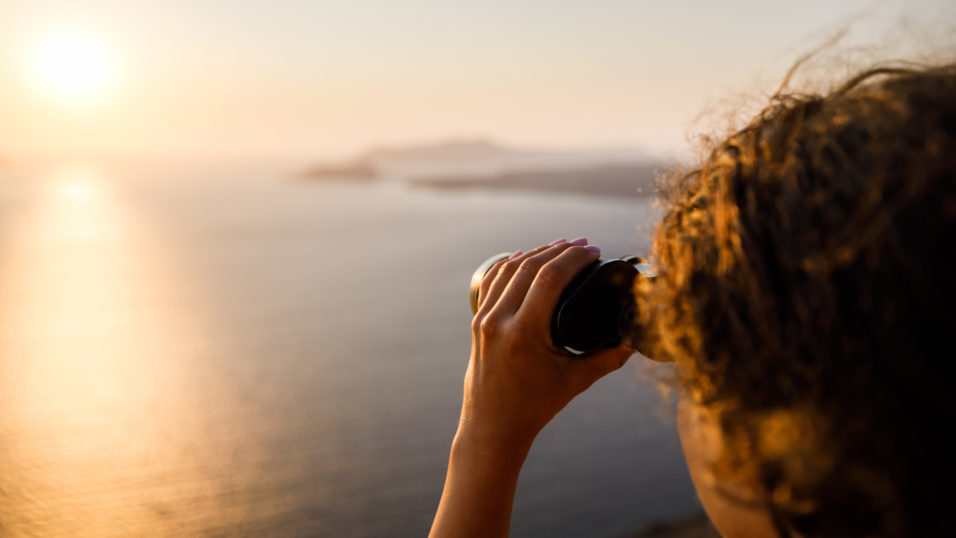 A woman looks through binoculars over water toward the setting sun.