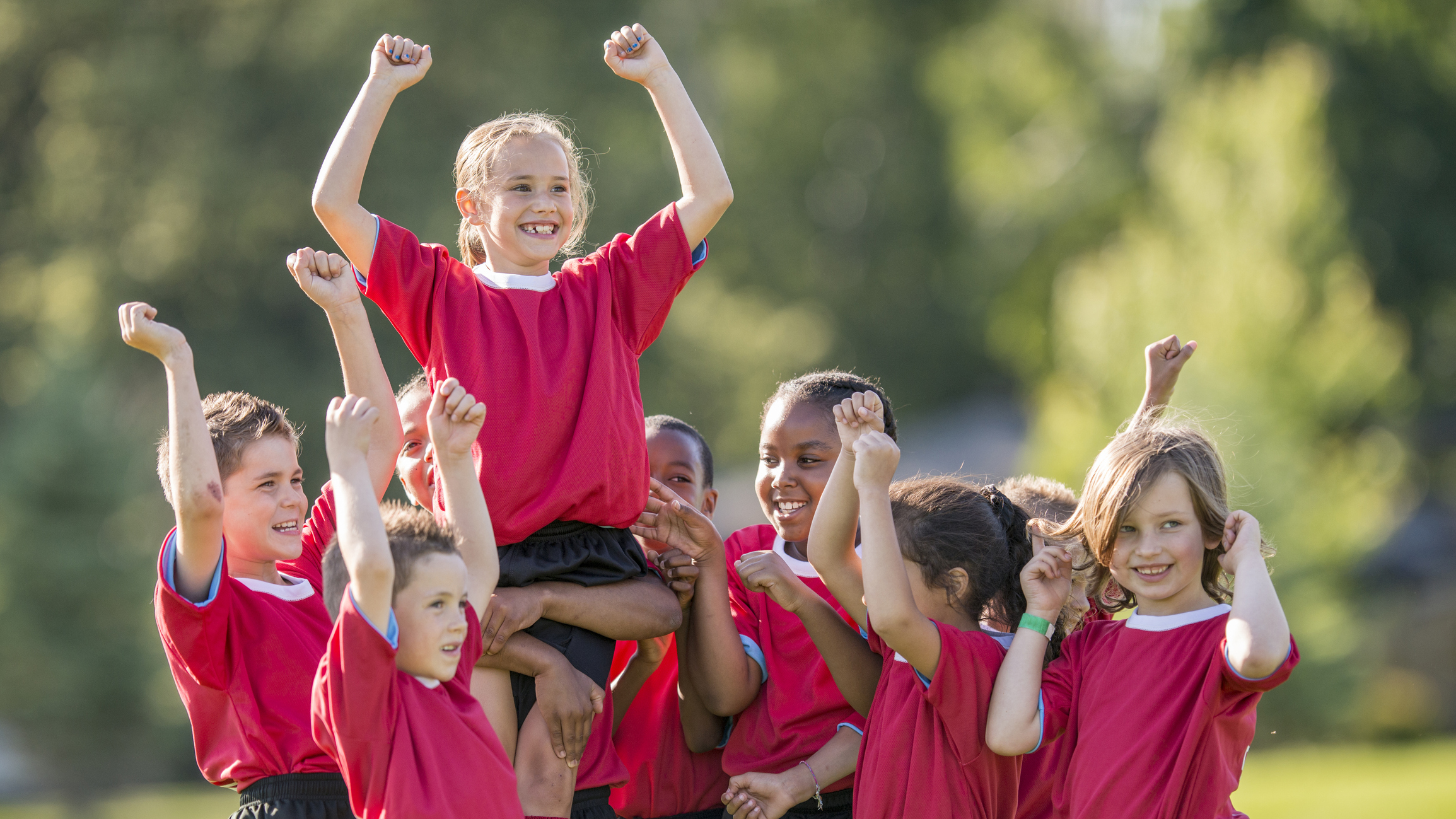 A group of elementary age children are lifting up on of their teammates for scoring the winning goal. The children are cheering and shouting victoriously.