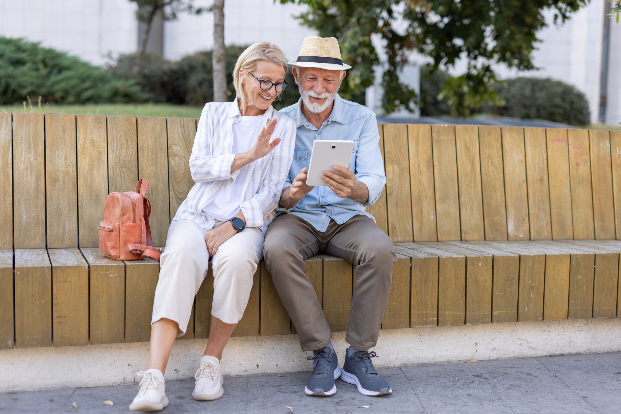 An elderly couple cheerfully uses a tablet together while sitting on a wooden bench in a sunny outdoor park, showcasing joy, companionship, and the embrace of digital life.