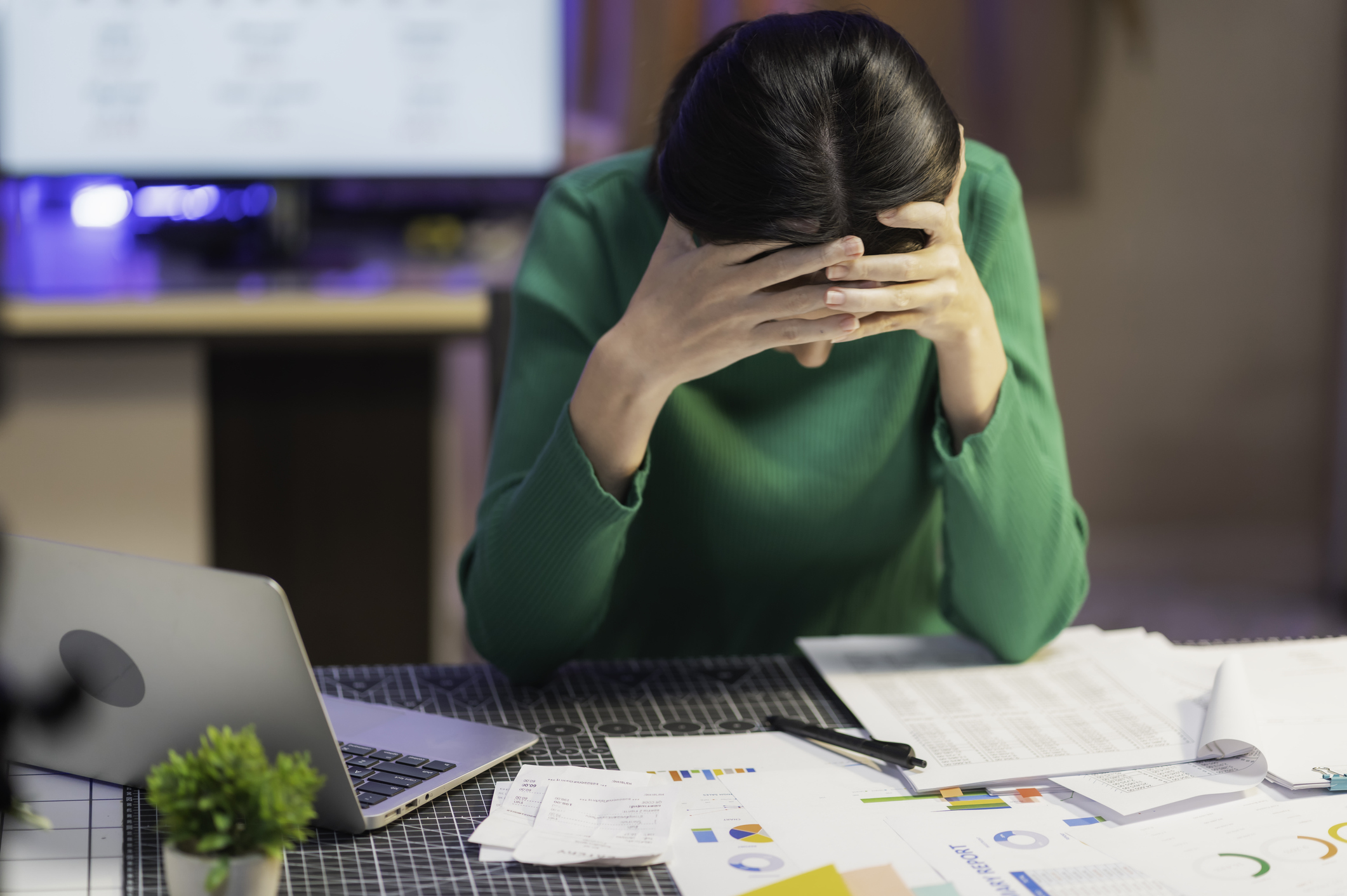 Stressed woman sitting at desk with laptop and financial documents, holding head while reviewing expenses and reports.