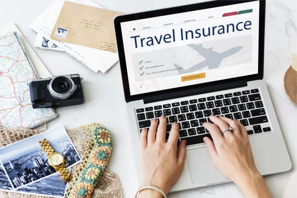 A person types on a laptop displaying a travel insurance website. Nearby are maps, a camera, postcards, photos, a woven hat, and a bracelet, suggesting travel planning.