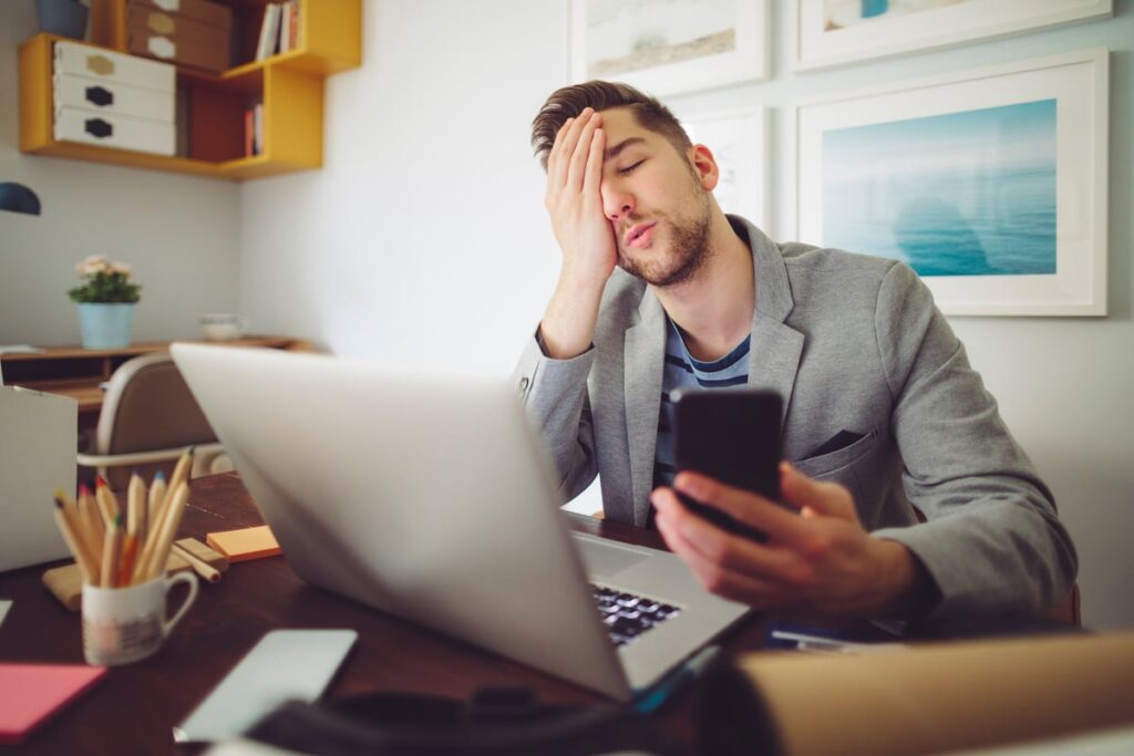 An investor sitting at a desk in front of a computer and holding a cell phone expresses regret.
