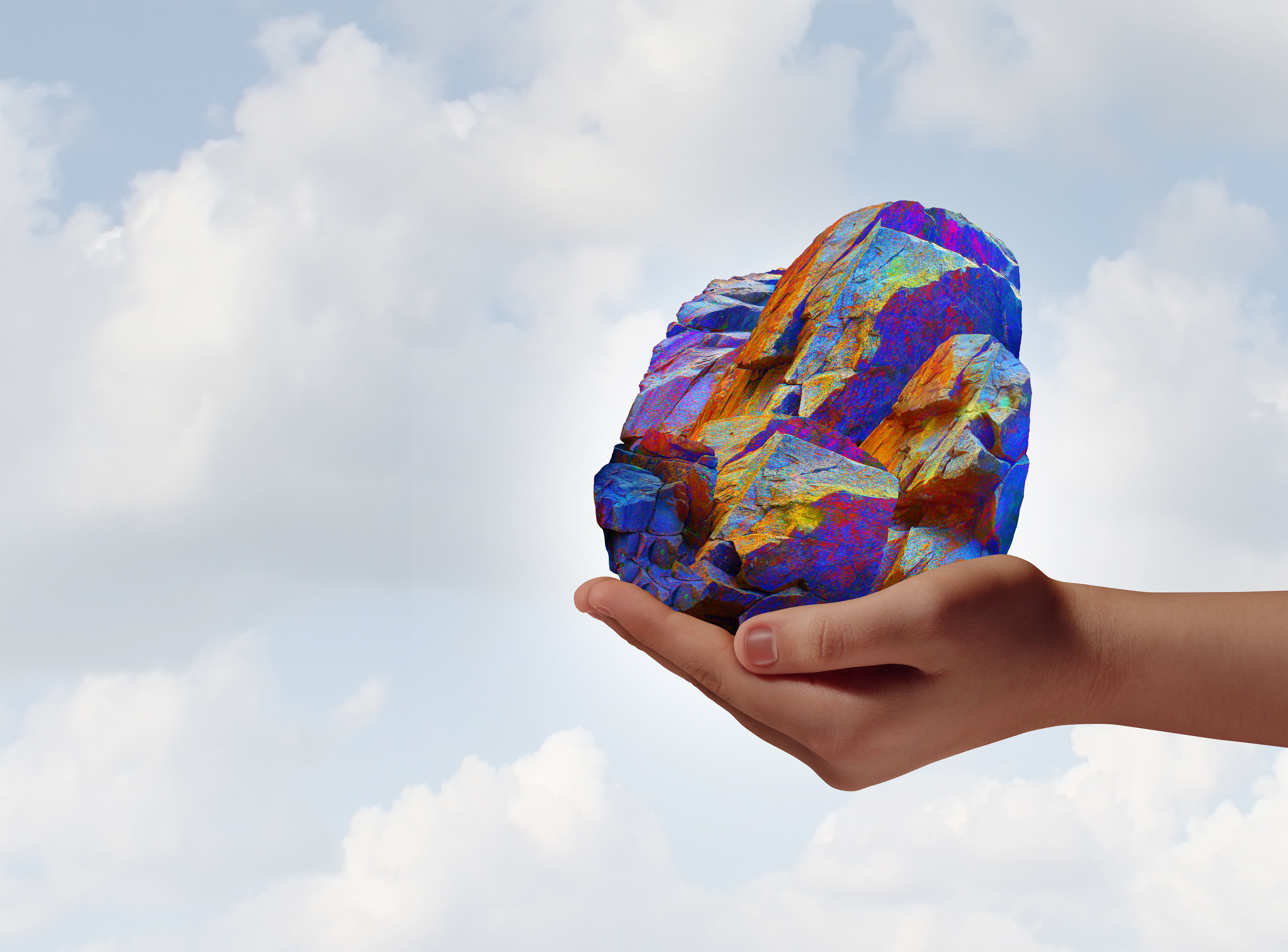 A hand holds up a colorful rock meant to represent rare-earth elements against the backdrop of a cloudy sky.