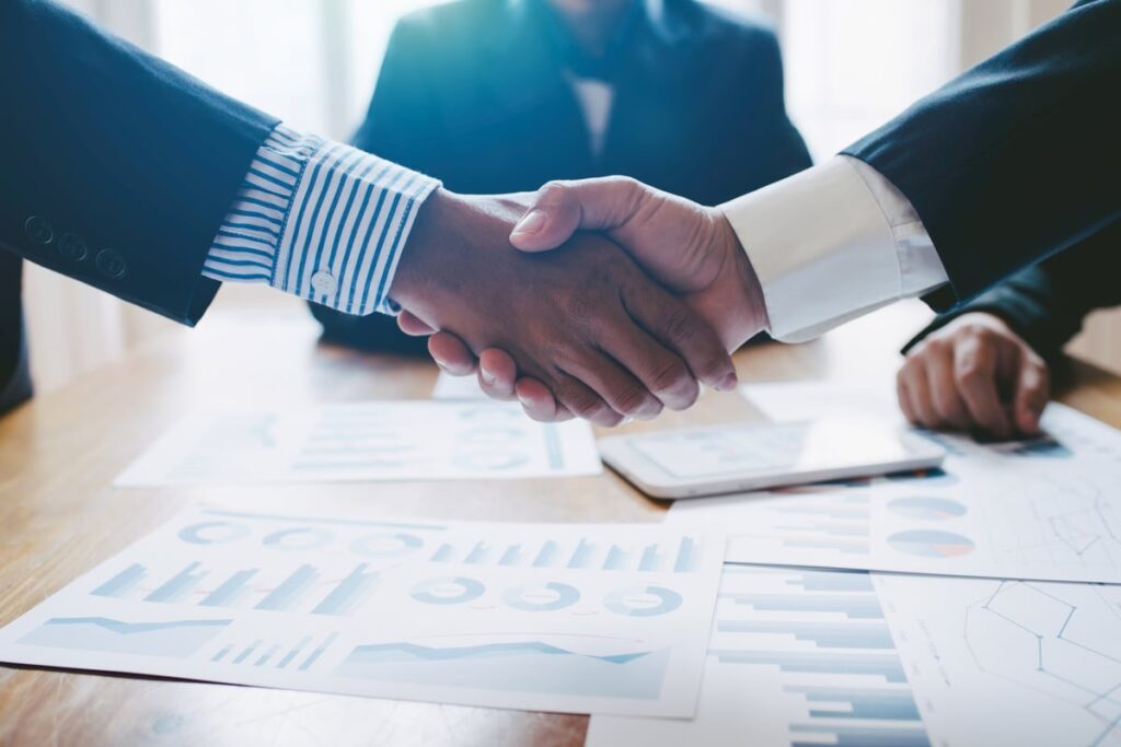 Two people shaking hands over a table full of diagrams and contracts.
