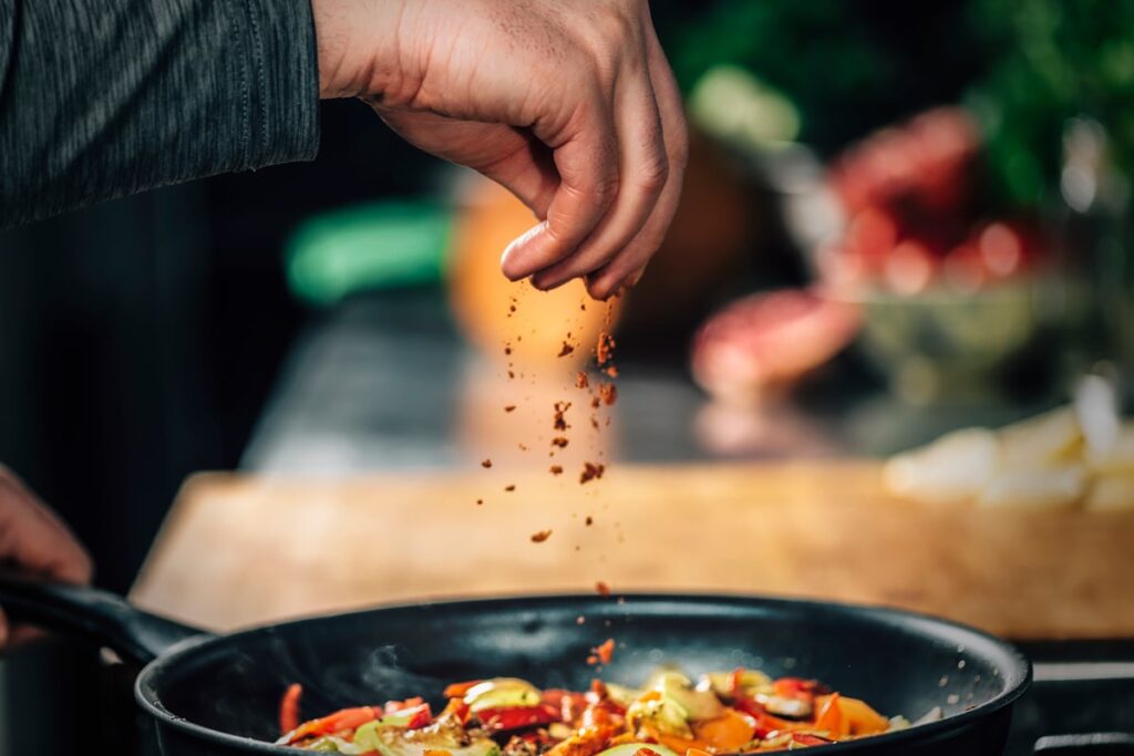 Person adding seasoning to pan of food.