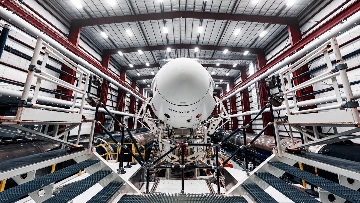 SpaceX rocket capsule in a brightly lit assembly hangar surrounded by platforms and support structures
