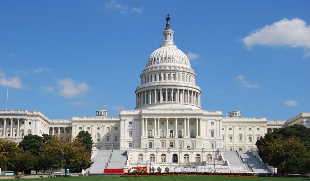 An eye-level, slightly upward-angled, daytime shot of the U.S. Capitol Building in Washington D.C. under a clear blue sky with sparse white clouds. The iconic white dome, topped with the Statue of Freedom, dominates the center of the frame, surrounded by its intricate columns and architectural details. The building's white marble facade extends horizontally, flanked by lush green trees on both sides, with more distant buildings partially visible. In the foreground, scaffolding and construction equipment are visible on the steps leading up to the Capitol, suggesting ongoing work. This image visually represents the "Congress" mentioned in the article, specifically the legislative body responsible for the "Big Beautiful Bill" that eliminates federal Grad PLUS loans, impacting graduate students' ability to afford professional programs like law and medicine.