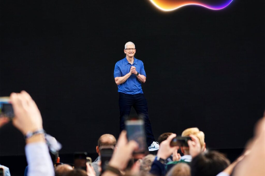 Tim Cook speaking on stage with a crowd of people taking photos and holding smartphones.