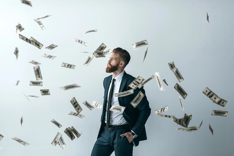 Bearded man in a dark business suit standing confidently with his hand in his pocket while US dollar bills rain down around him against a gray background. This illustrates the potential for wealth accumulation and passive income through high-yield savings accounts, which are currently offering interest rates up to 4.15% APY as of September 2026, allowing savers to maximize returns despite fluctuating market conditions. Source: The College Investor