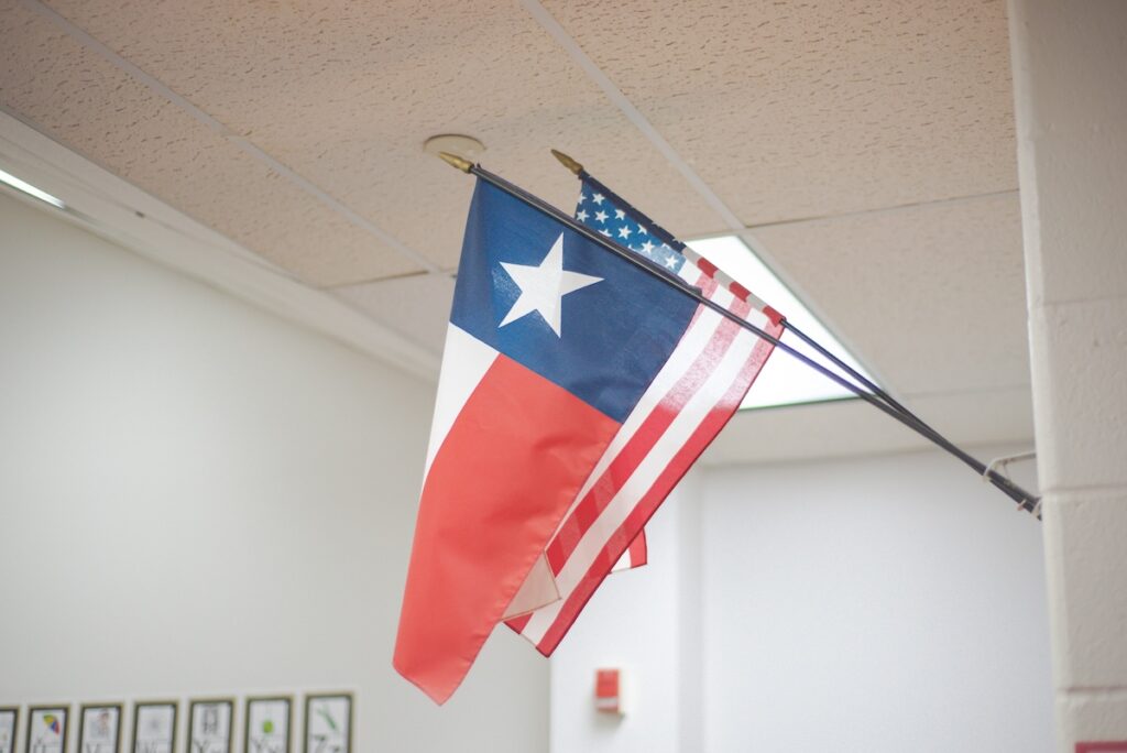 School in Dallas Fort Worth highlights Texas flag angled beside national banner on white wall.